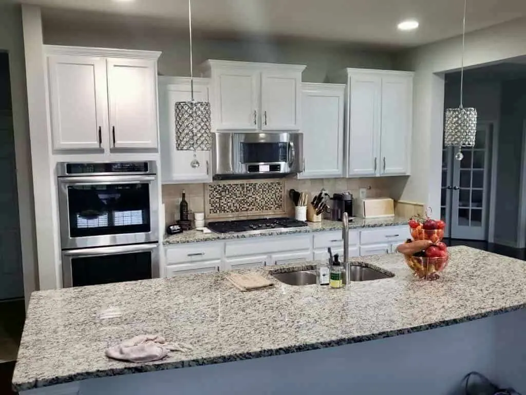 A brightly lit, modern kitchen in Geist showing the completed results of painting kitchen cabinets white after a professional refinishing by the guy painting team. The image features a large central island with a granite countertop and a bowl of fresh fruit, leading to the main cabinetry wall. The shaker-style upper and lower cabinets are finished in a clean, durable white that coordinates perfectly with the stainless steel double ovens, microwave, and refrigerator. A detailed mosaic tile backsplash behind the gas cooktop and elegant crystal pendant lighting complete the sophisticated look of this kitchen transformation by guy painting.