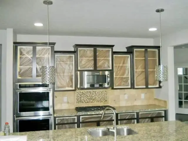 A professional "before" documentation of a high-end Geist kitchen renovation, capturing the essential preparation steps for painting kitchen cabinets white before the final coat. The image shows a large kitchen island and perimeter cabinetry originally in a medium wood tone, now being meticulously prepped by the guy painting team. The countertops and floors are completely covered in protective brown paper and plastic, while the backsplash is carefully masked to ensure a clean, sharp finish. This thorough prep work by guy painting is the foundation for transforming this traditional space into a bright, modern white kitchen.