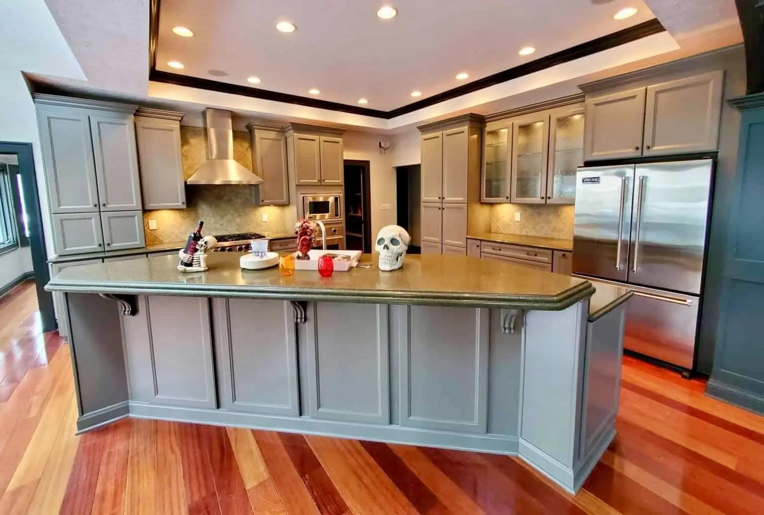 A polished "After" view of a high-end kitchen transformation, showcasing the expert results of painting old kitchen cabinets After by the guy painting crew. The cabinetry, including the large, multi-angled kitchen island and full-height pantry units, is finished in a smooth, professional coat of Gauntlet Gray. The sophisticated dark gray tone creates a modern, upscale feel that contrasts beautifully with the warm, glossy hardwood floors. This comprehensive refinishing project by guy painting features integrated stainless steel appliances, a sleek range hood, and specialized glass-front upper cabinets with interior lighting, all unified by the precision paint application.