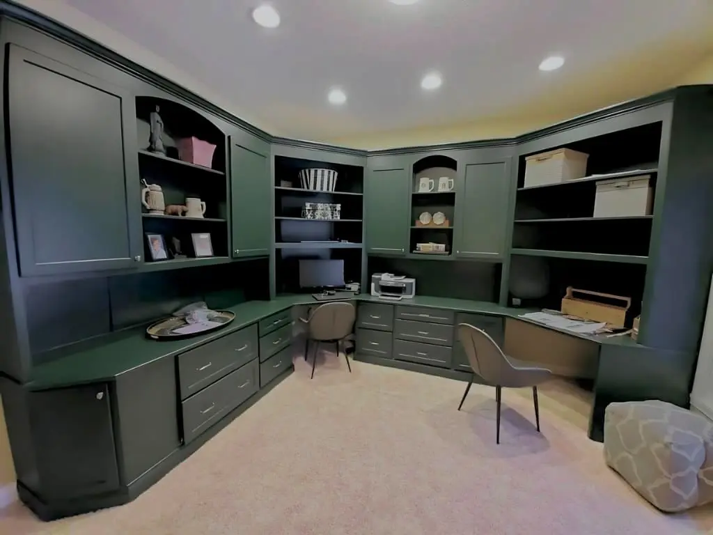 A large, wrap-around home office featuring a custom built-in desk and shelving unit that has received a professional cabinet painting indianapolis transformation. The cabinetry is finished in a deep, sophisticated forest green with a smooth, durable sheen. The unit includes multiple drawers with sleek silver pulls, open shelving for organization and decor, and a large corner desk area. The dark cabinetry provides a striking contrast against the light-colored plush carpeting and soft wall tones, demonstrating the high-quality craftsmanship and modern aesthetic delivered by the guy painting team in Geist.