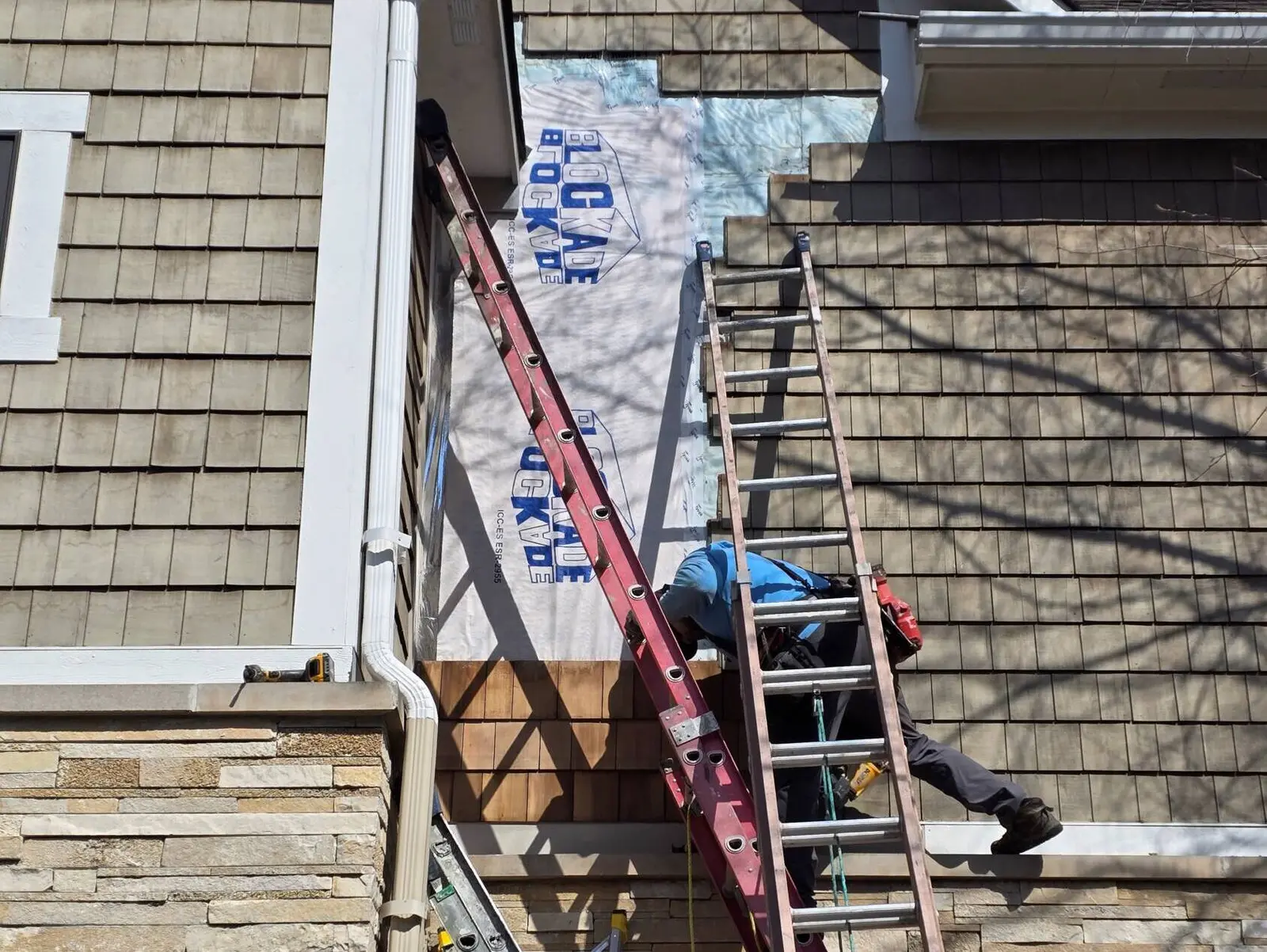 A professional technician on a tall red ladder performs detailed maintenance on a residential home's cedar shingle siding. This project highlights the thorough exterior painting and repair services available in geist. Guy painting focuses on essential structural preparation, including the installation of protective house wrap and shingle replacement, to ensure a durable and high-quality finish for every residential property.