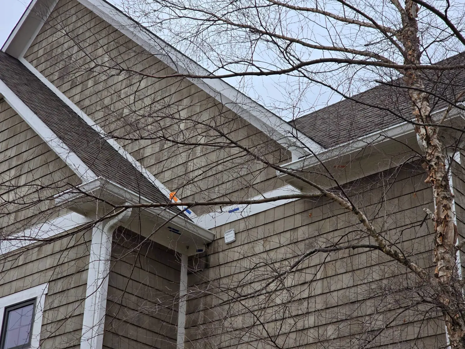 A close-up view of a residential home's cedar shingle siding and white gutter system, showing blue and orange tape markers used for identifying necessary maintenance areas. This detailed inspection is a core part of our exterior painting and repair services in geist. Guy painting carefully assesses the condition of the exterior wood and trim to ensure all issues are addressed before a fresh, protective finish is applied.